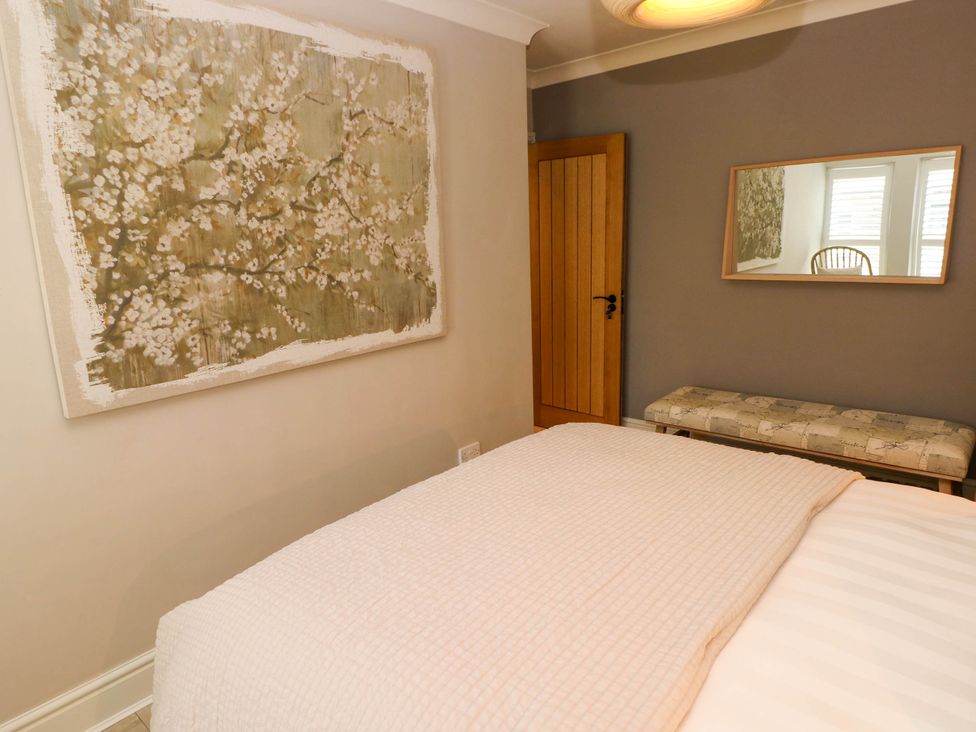 A bedroom with a bed covered with a white blanket a wooden door a floral painting a bench and a mirror on the wall at The Loft in Ystradgynlais