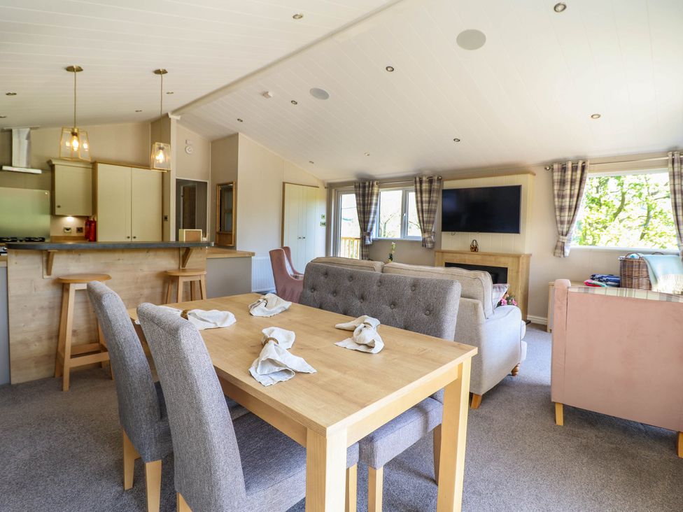 A dining area with a wooden table and gray chairs next to a living room with sofas a TV and windows at The Owlet in Dolwen near Llanidloes