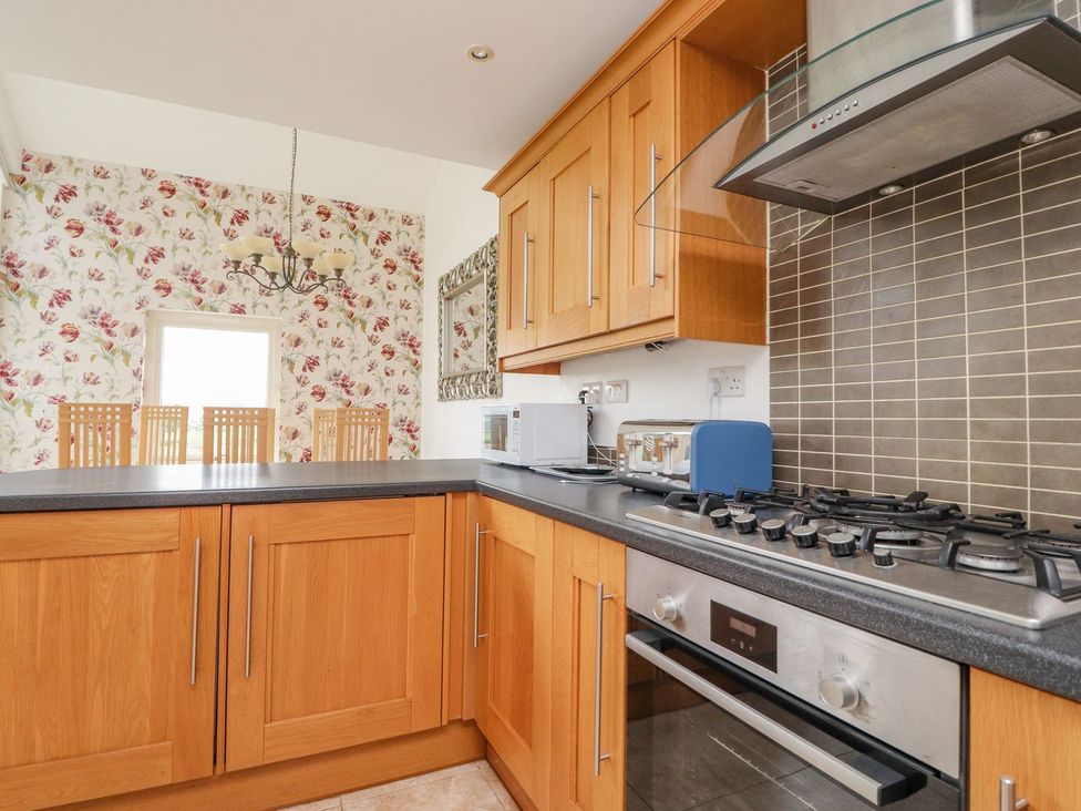 A kitchen with wooden cabinets a countertop a gas stove a microwave toaster dining chairs and floral wallpaper at High View Meadows in Kirkby Lonsdale