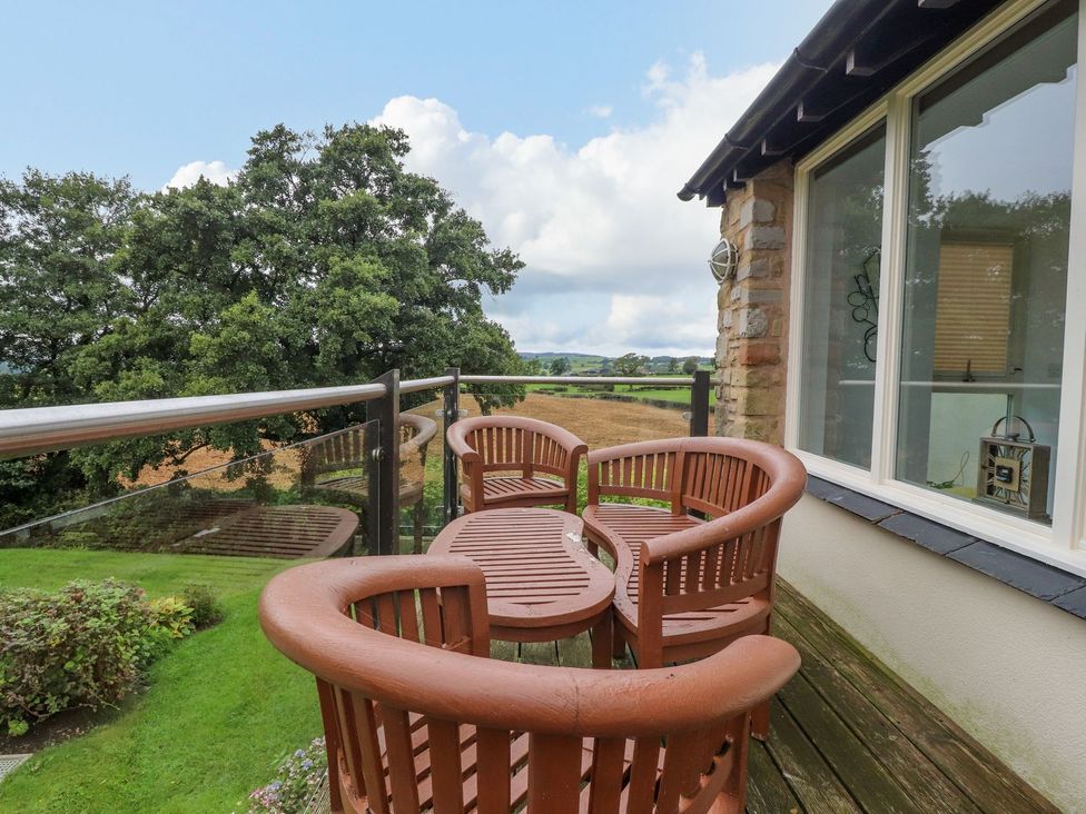An outdoor patio with wooden chairs and a table overlooking trees and fields at High View Meadows in Kirkby Lonsdale