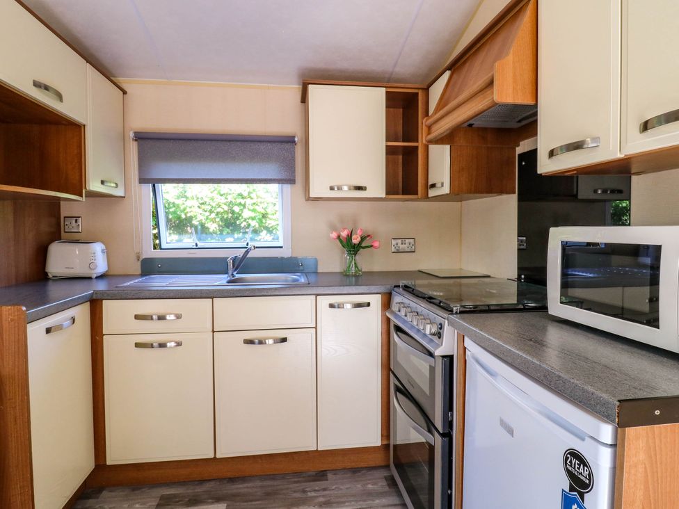 A kitchen with cream cabinets a gray countertop a window with a blind a stove a microwave a refrigerator a toaster and a vase with flowers at Caravan 941 in Clacton-On-Sea