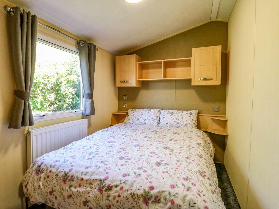 A bedroom with a bed floral bedding a window with curtains and wall cabinets at Caravan 941 in Clacton-On-Sea