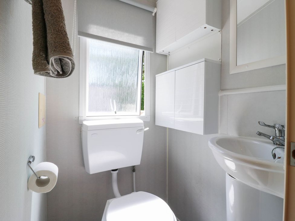 A small bathroom with a toilet, sink, white cabinet, rolled towel, toilet paper holder, and frosted window at Caravan 941 in Clacton-On-Sea