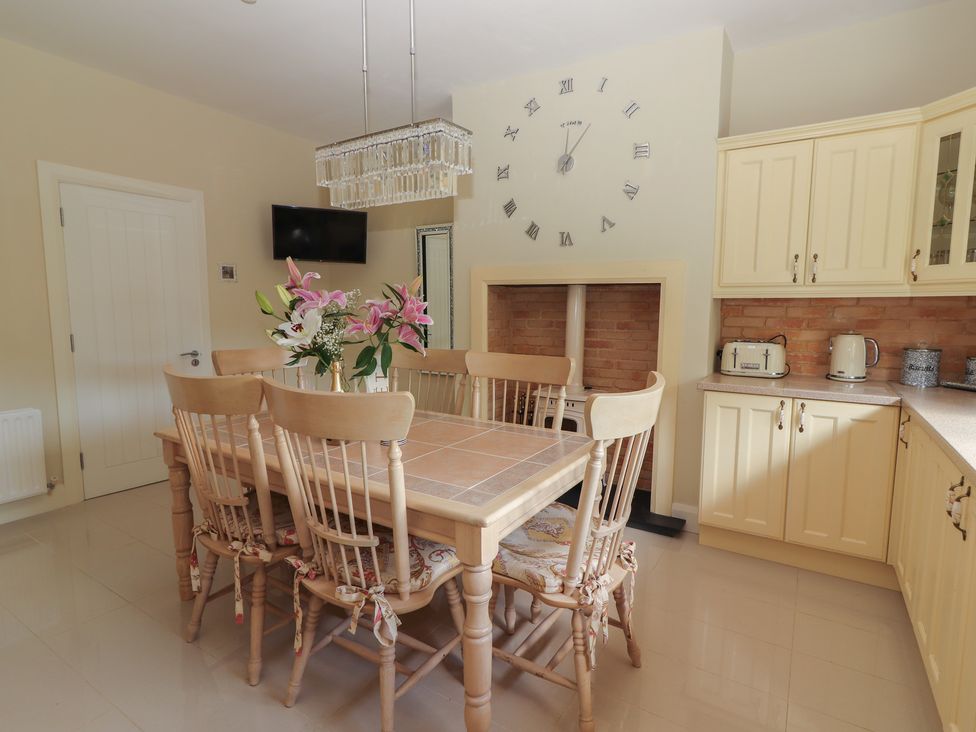 A kitchen dining area with a wooden table and six chairs a vase of flowers a wall clock a small tv and cream cabinets at Killelton Cottage Camp County Kerry