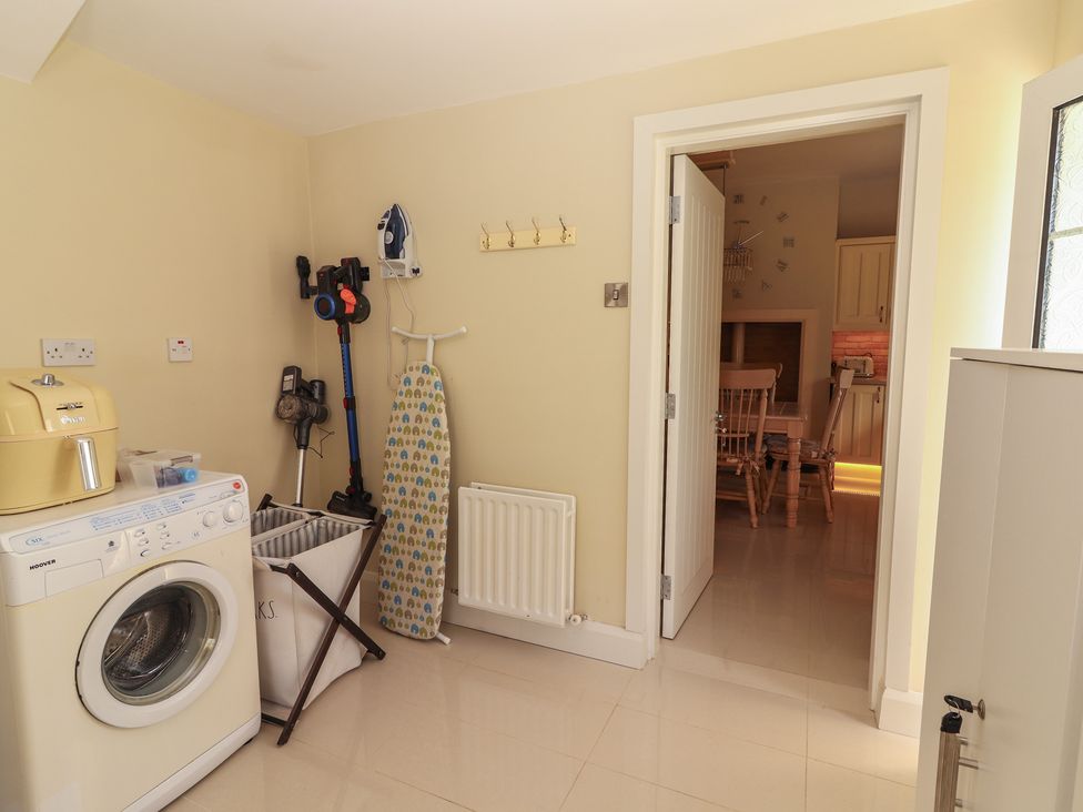 A utility room with a washing machine ironing board laundry basket and vacuum cleaners next to a doorway leading to a kitchen with wooden chairs at Killelton Cottage in Camp County Kerry