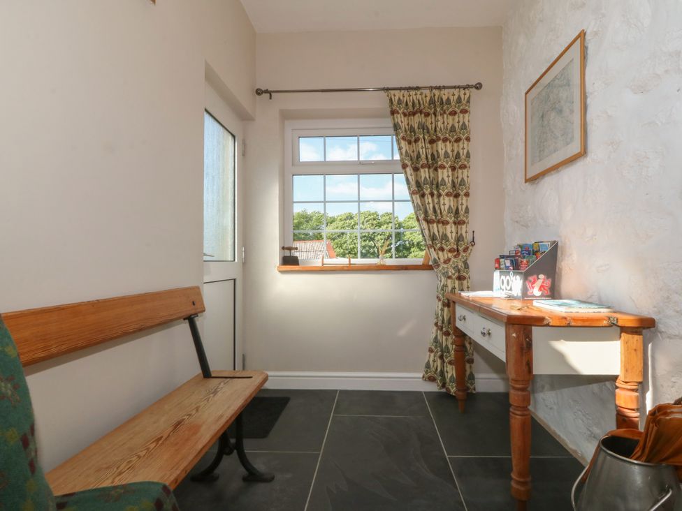 A small entryway with a wooden bench, a window with patterned curtain, and a wooden table with assorted items at Glasfryn in Llandwrog