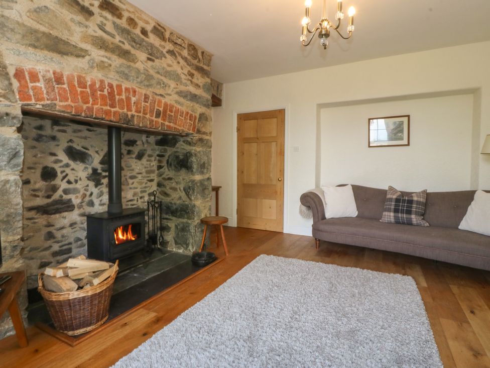 A living room with a stone fireplace and wood stove a basket of firewood a gray sofa and a wooden door at Glasfryn in Llandwrog
