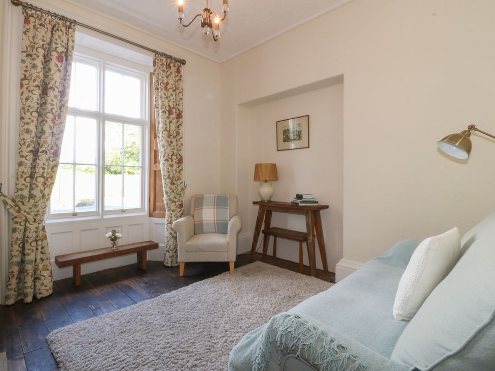 A sitting room with armchair beside a window with floral curtains and a wooden console table with lamp and books at Glasfryn in Llandwrog