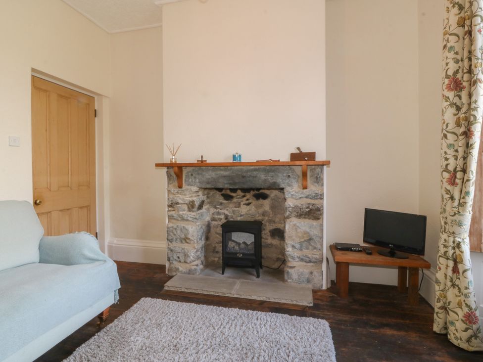 A room with a stone fireplace, a small electric heater, a wooden door, a light blue sofa, a wooden table with a television, and floral curtains at Glasfryn in Llandwrog