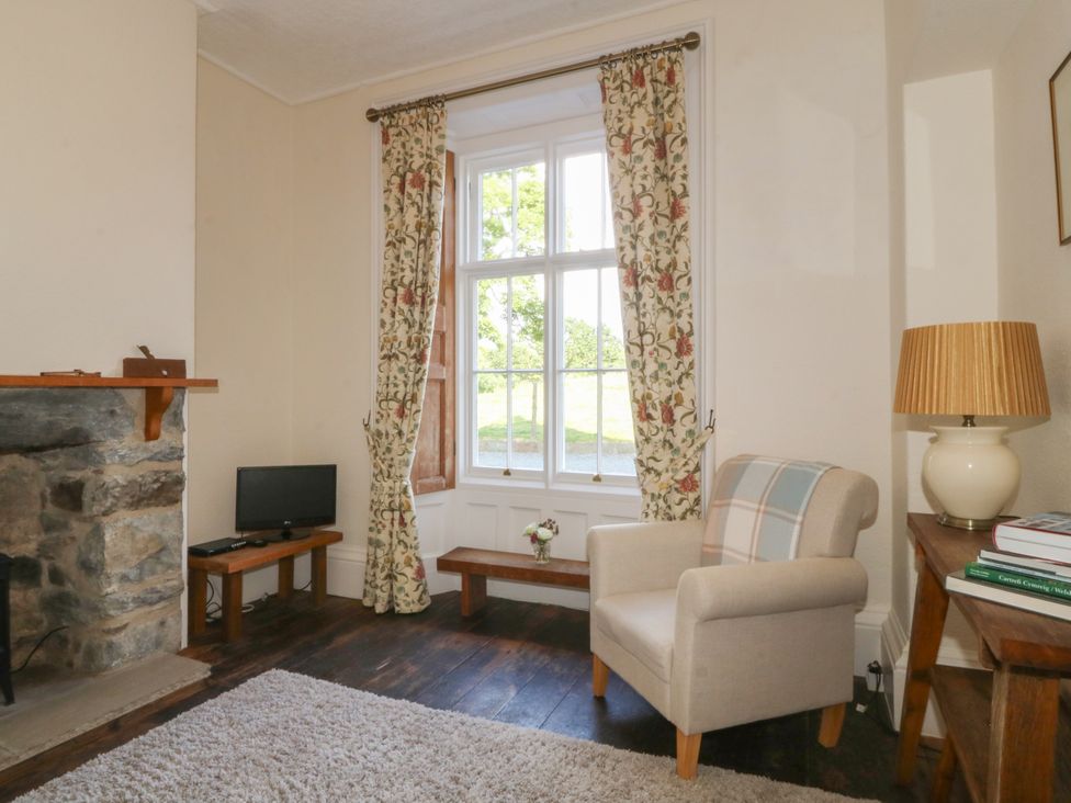 A sitting room with a stone fireplace a beige armchair and a window with floral curtains at Glasfryn in Llandwrog