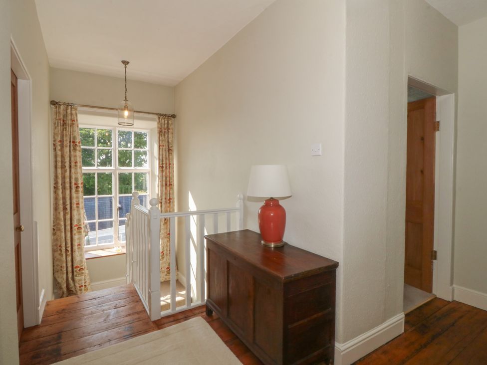 A hallway with wooden floor a wooden cabinet with a lamp white window with curtains and a staircase at Glasfryn in Llandwrog