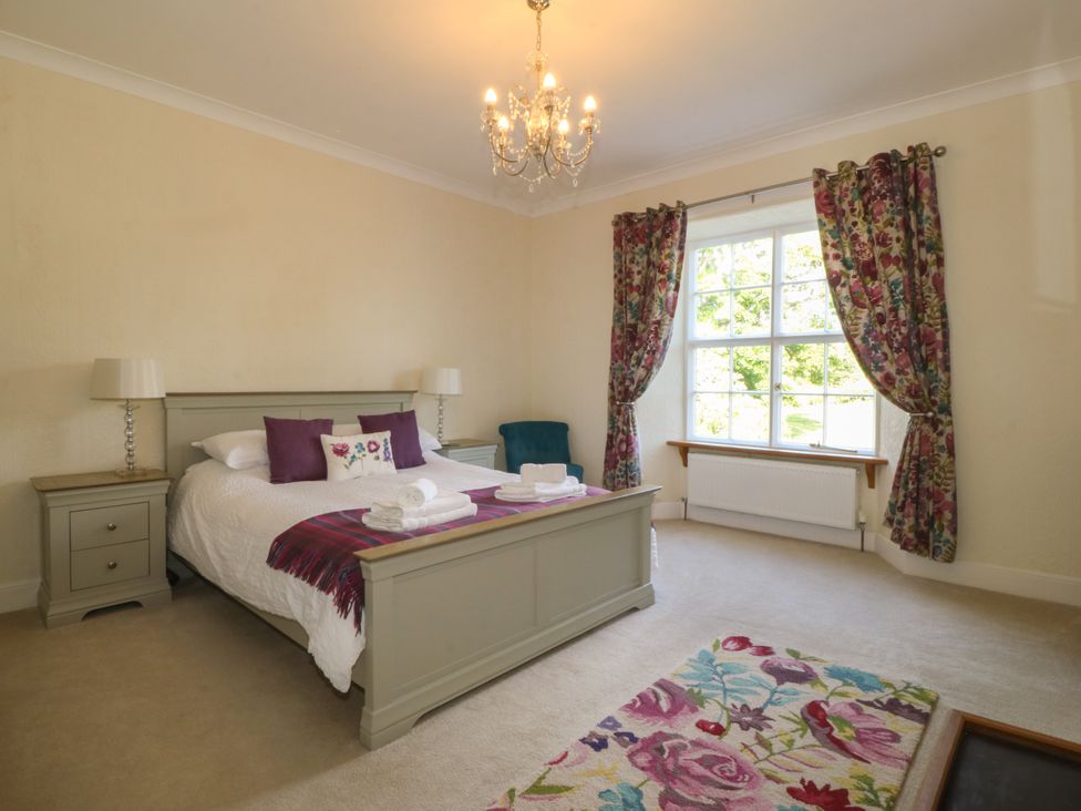 A bedroom with a large bed two nightstands with lamps a window with floral curtains and a floral rug at Glasfryn in Llandwrog