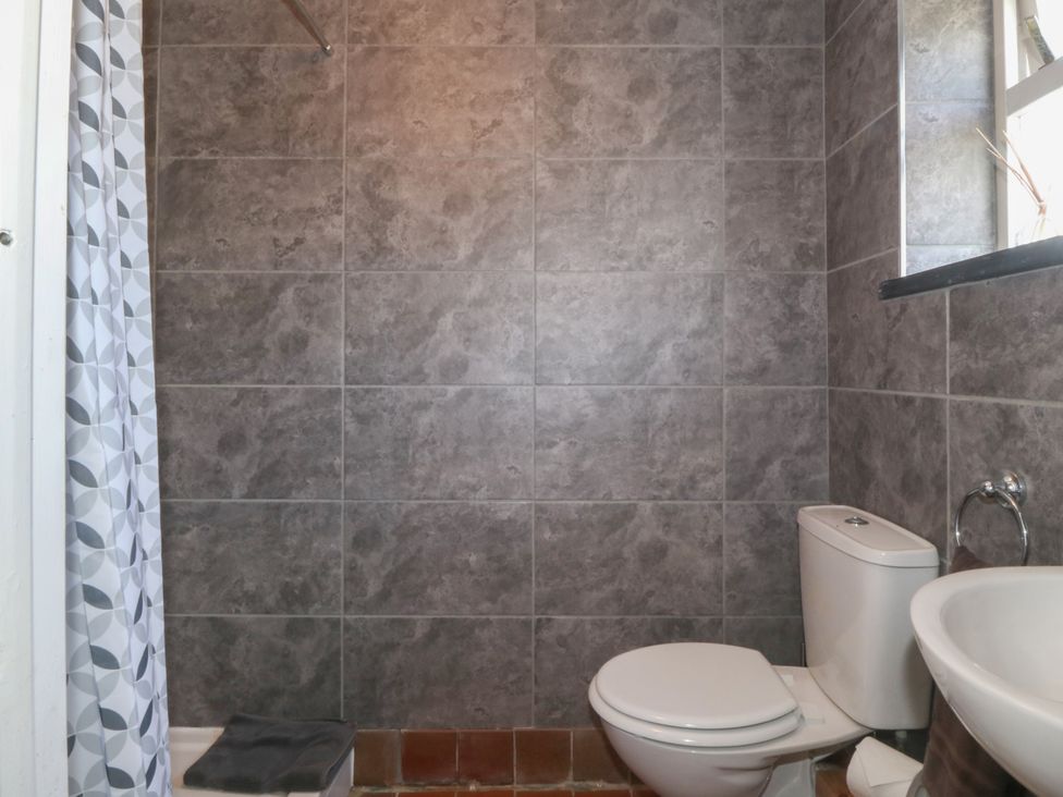 A bathroom with a toilet, sink, shower area with a patterned curtain, and grey tiled walls at Glasfryn in Llandwrog