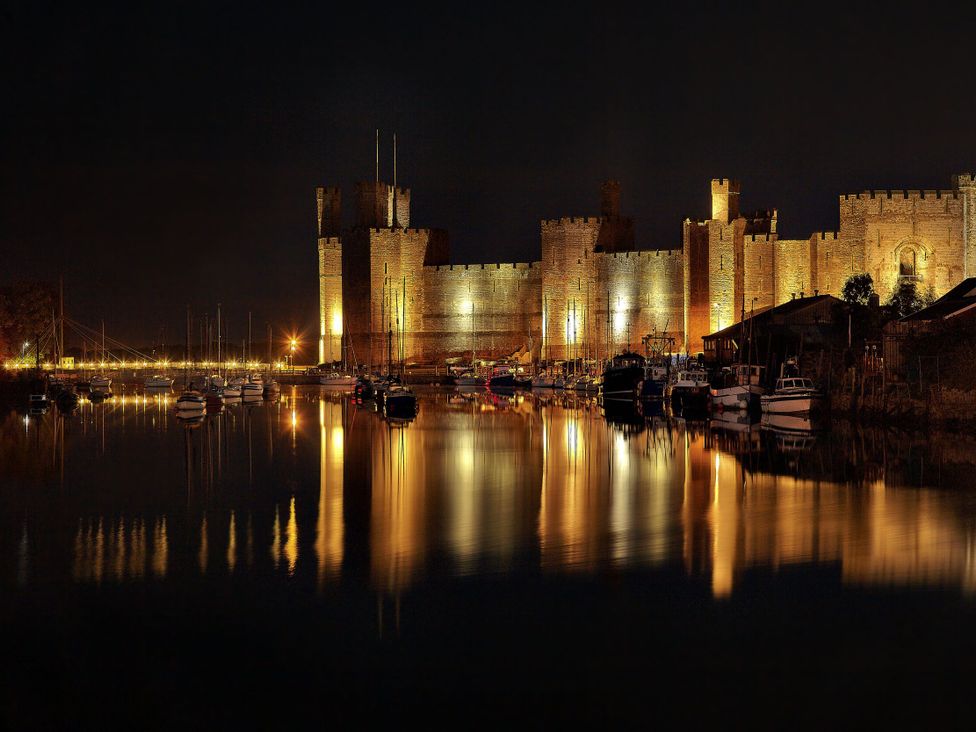 A castle illuminated at night beside a river with boats at Caernarfon Castle in Caernarfon