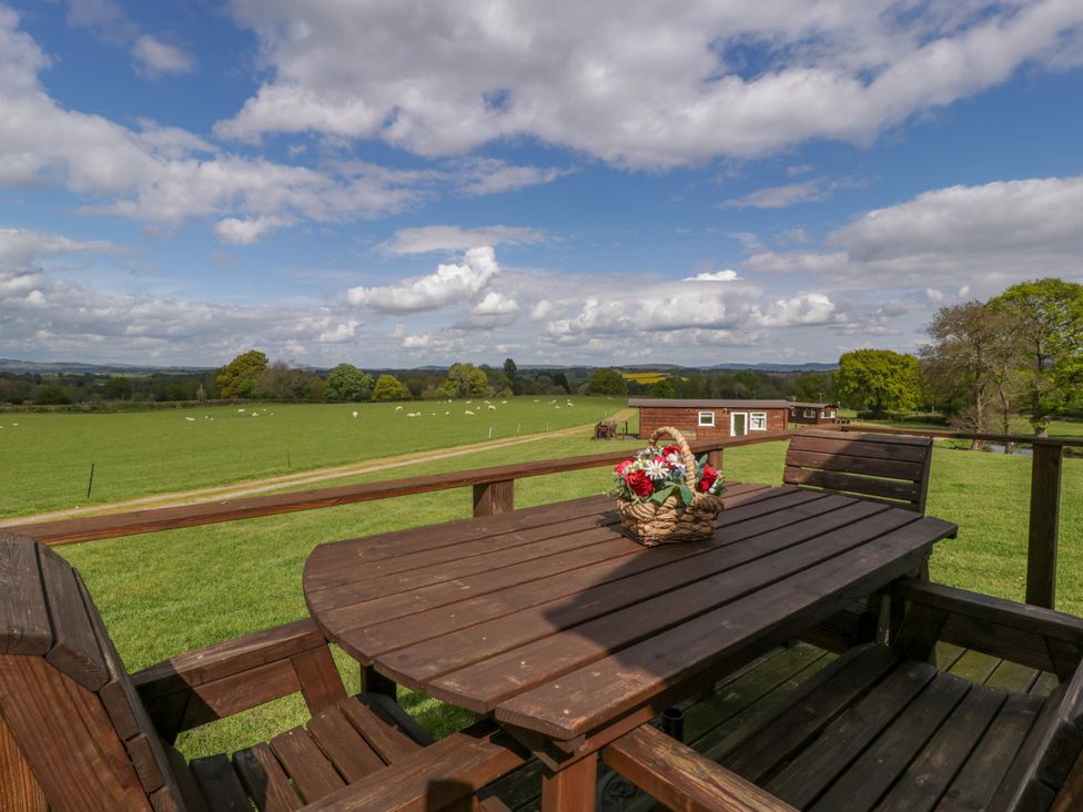 A wooden outdoor table and chairs with a basket of flowers on the table overlooking a green field with sheep and a distant building at Twilight in Raglan