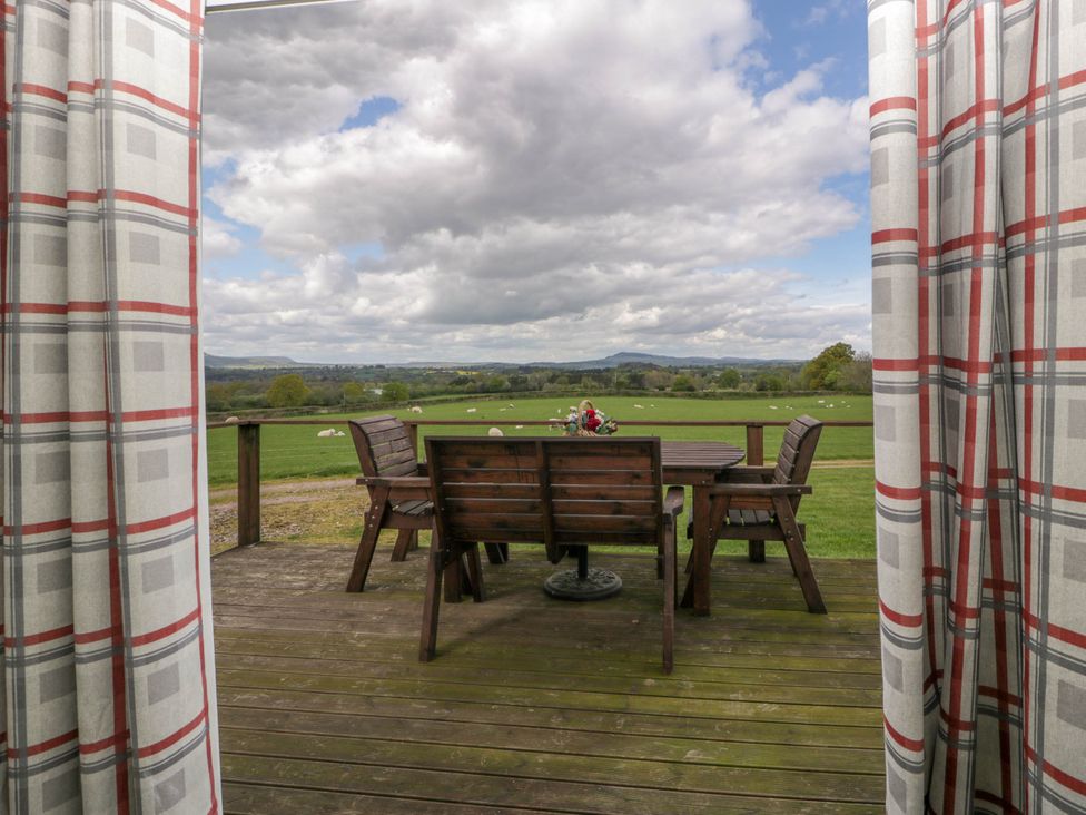 A wooden deck with a table and chairs and a basket of flowers overlooking a green field with sheep and hills in the distance at Twilight in Raglan