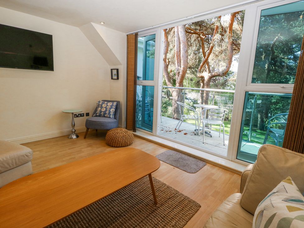 A living room with a sofa, wooden coffee table, armchair, side table, wall-mounted TV, and glass doors opening to a balcony with metal chairs and table at 6 Red Sails in Sandbanks