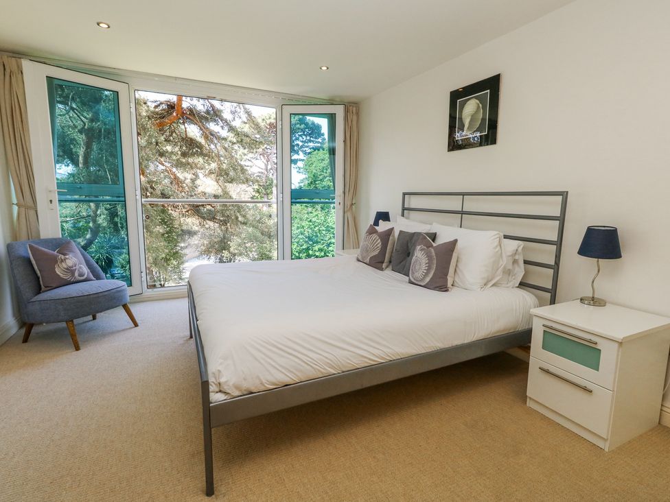 A bedroom with a bed with pillows a bedside table with a lamp a chair and large windows at 6 Red Sails in Sandbanks