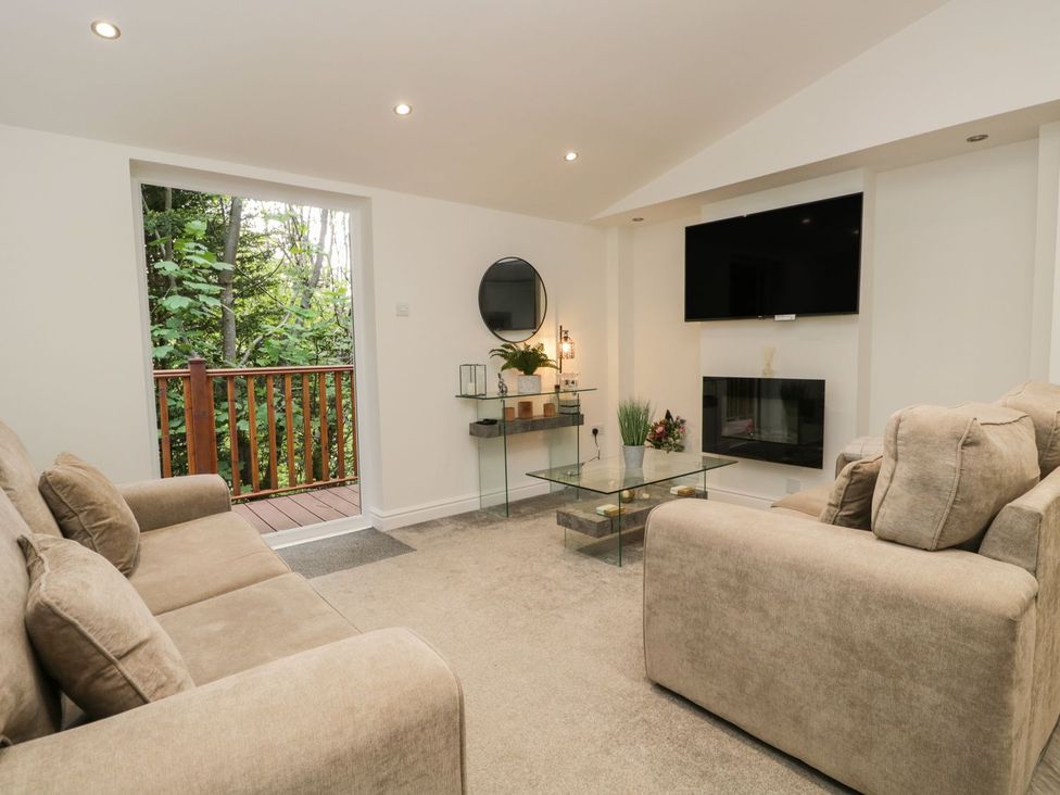 A living room with two beige sofas a glass coffee table a wall-mounted TV and a glass door opening to a wooden balcony at Mere Brook Lodge in White Cross Bay