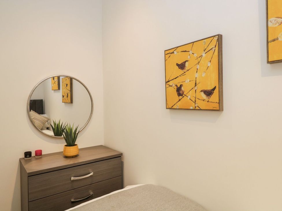 A bedroom corner with a wooden dresser holding a plant in a yellow pot and two candles with a round mirror above it and yellow bird paintings on the wall at Mere Brook Lodge White Cross Bay