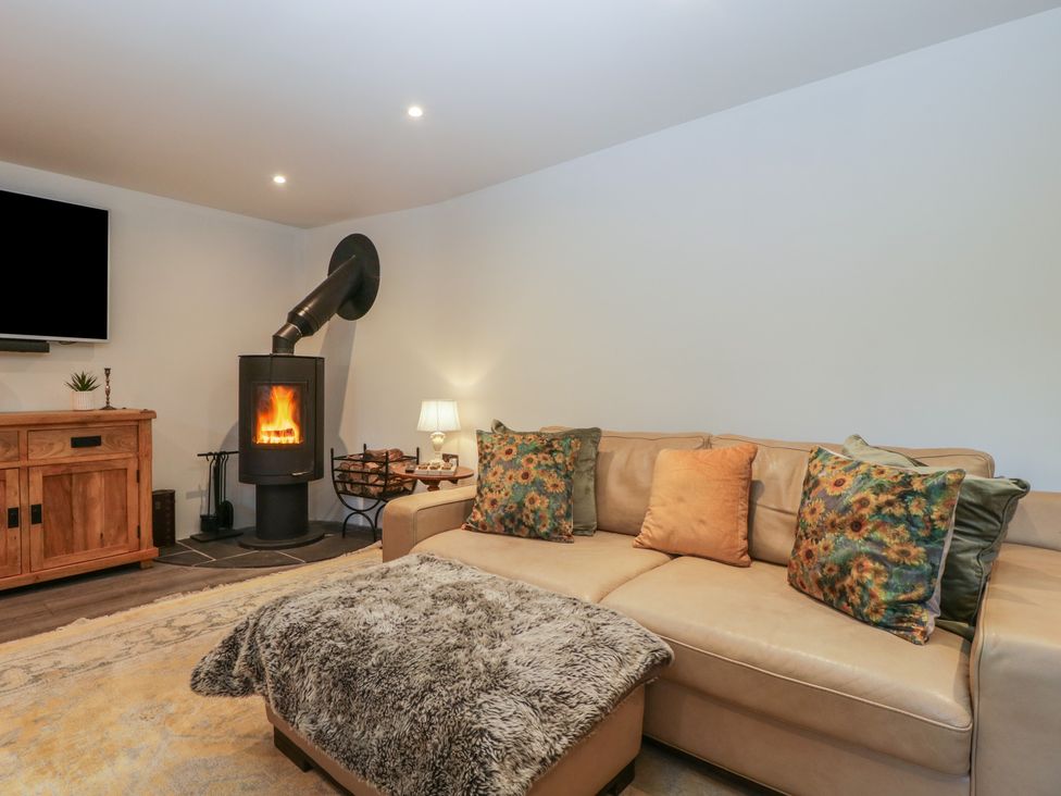 A living room with a beige couch with flower and solid color cushions a wood burning stove a wooden cabinet and a TV at Graig Cottage in Cwmcarn
