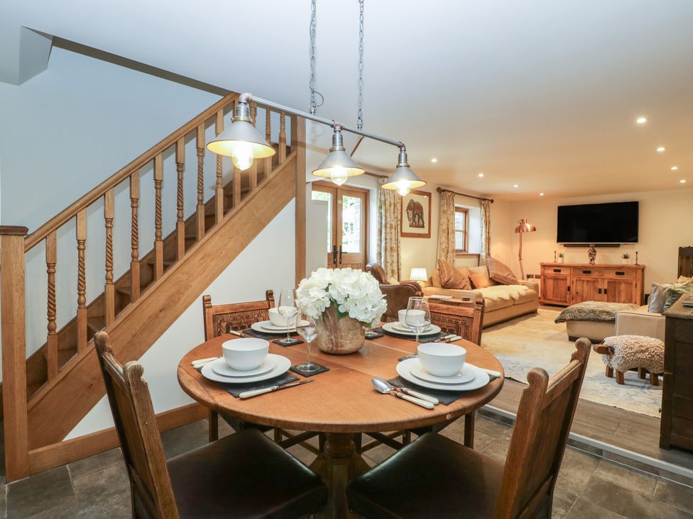 A dining area with a round table set for four near wooden stairs and a living room with sofas a television and a wooden cabinet at Graig Cottage in Cwmcarn