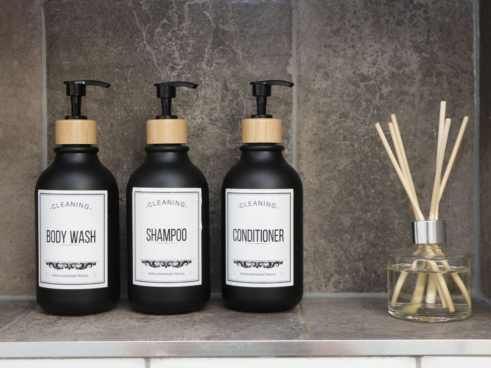 Three black bottles labeled body wash shampoo and conditioner next to a reed diffuser on a tiled surface