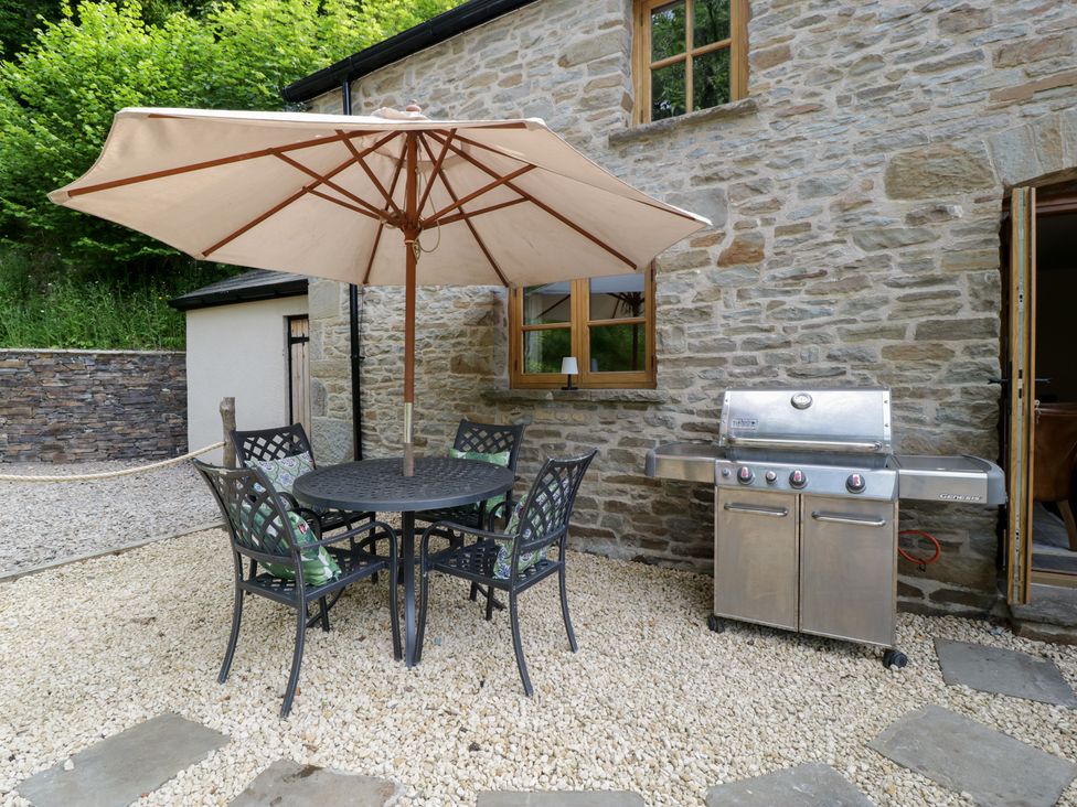 An outdoor patio with a round table and four chairs under a large umbrella next to a stainless steel barbecue at Graig Cottage in Cwmcarn