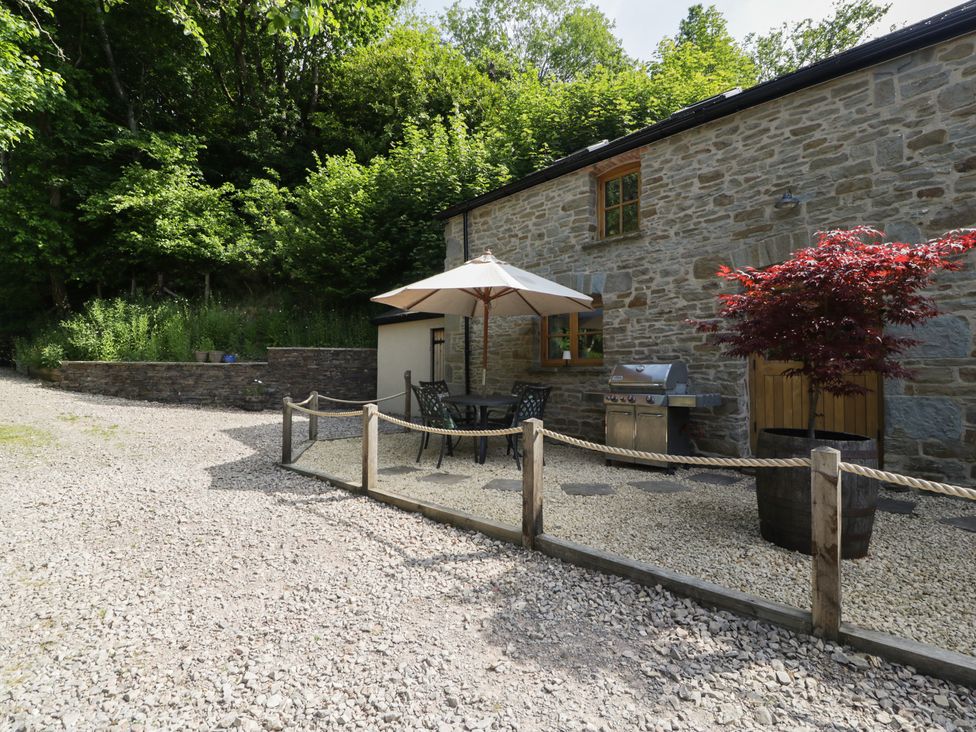 An outdoor patio area with a table umbrella chairs a barbecue and a tree in a planter by a stone building at Graig Cottage in Cwmcarn
