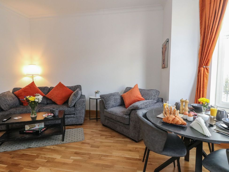 A living room with grey sofas and orange cushions a black coffee table with flowers and board games and a dining table with breakfast items at Lenas Corner in Dunoon