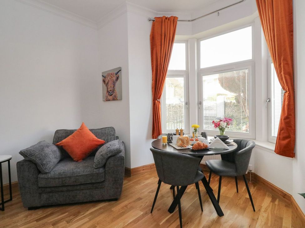 A dining table with breakfast items and gray chairs next to a gray armchair with an orange cushion and orange curtains at Lenas Corner in Dunoon