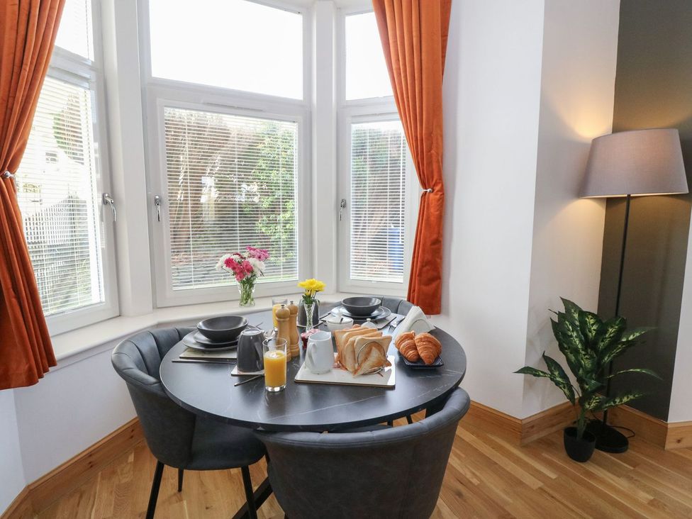 A dining area with a round table set for breakfast near bay windows with orange curtains at Lenas Corner in Dunoon
