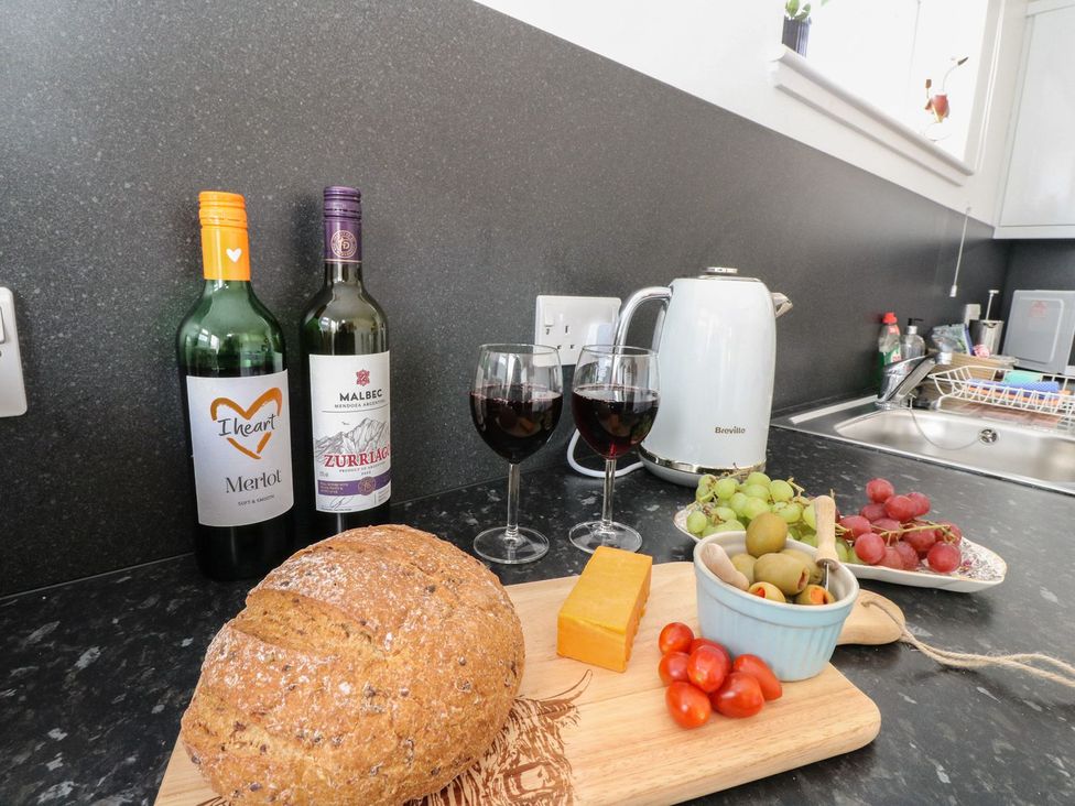 A kitchen counter with bread cheese olives tomatoes grapes two glasses of red wine two wine bottles and a white kettle at Lenas Corner in Dunoon