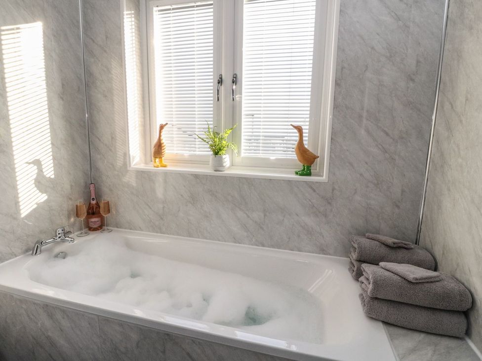 A bathroom with a bubble bath in a bathtub a window with blinds two duck figurines a plant two glasses and a bottle of pink drink and folded towels at Lenas Corner in Dunoon