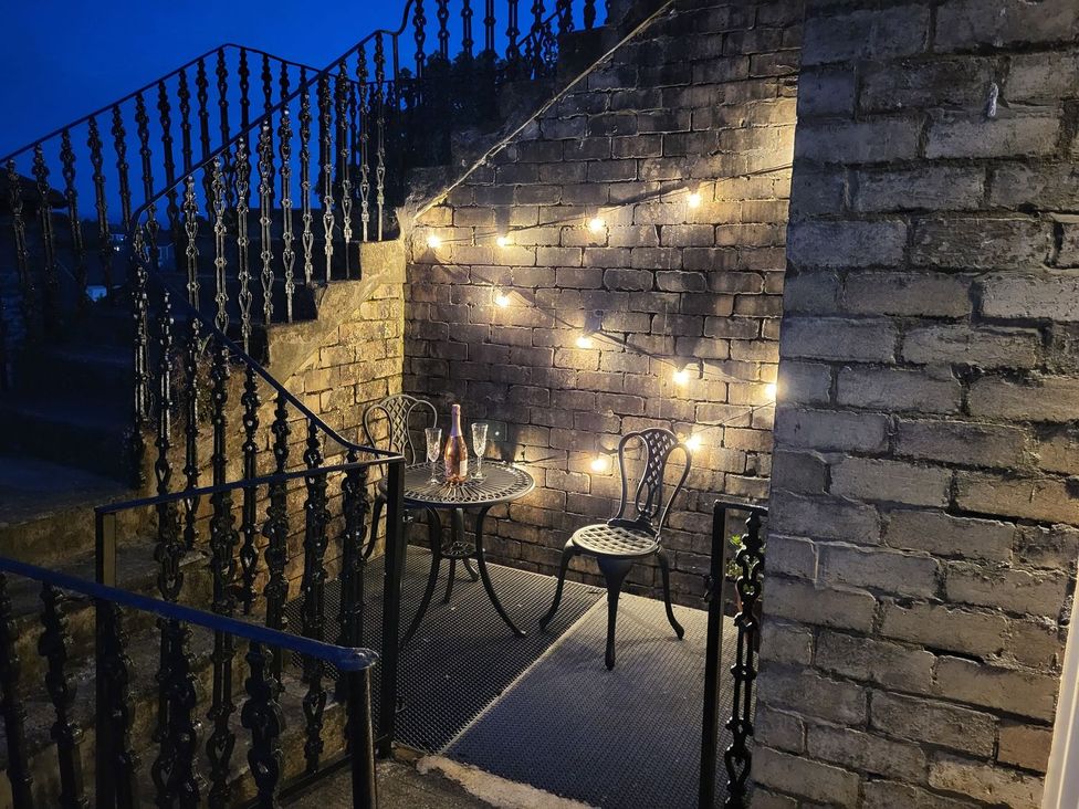 An outdoor seating area with a small round table and two chairs next to a brick wall with string lights at Lena's Corner in Dunoon