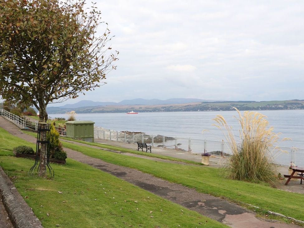 A grassy area with a tree path and bench next to a body of water at Lena's Corner in Dunoon