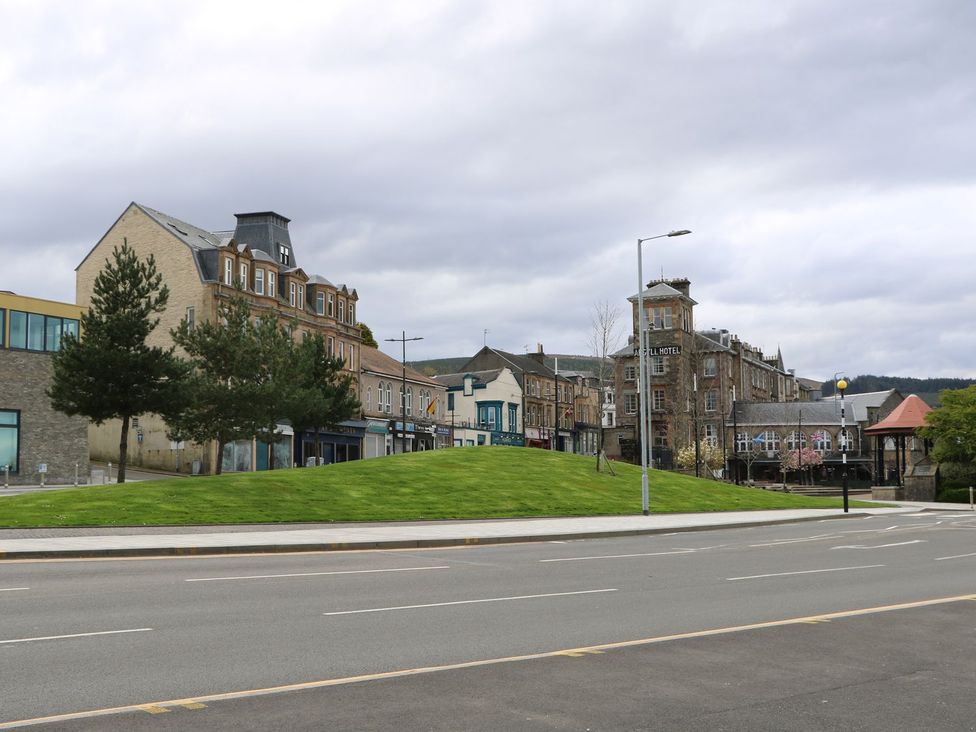 A street with a grassy mound and trees in front of buildings including a hotel and shops at Lenas Corner in Dunoon