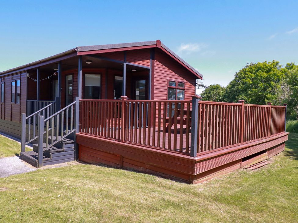 An exterior view of a wooden lodge with a fenced porch and steps leading to a grassy area at Lodge 9- Lamorna in Tregolds near St Merryn