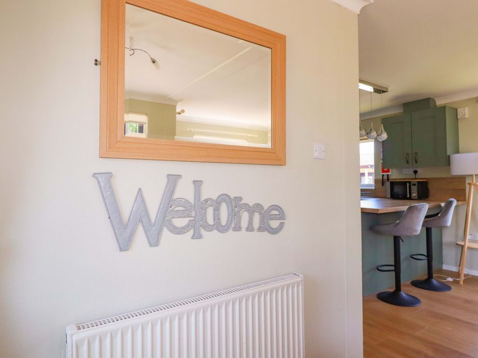 A wall with a rectangular wooden mirror and metal welcome sign above a white radiator with a kitchen area having two bar stools at Lodge 9- Lamorna in Tregolds near St Merryn