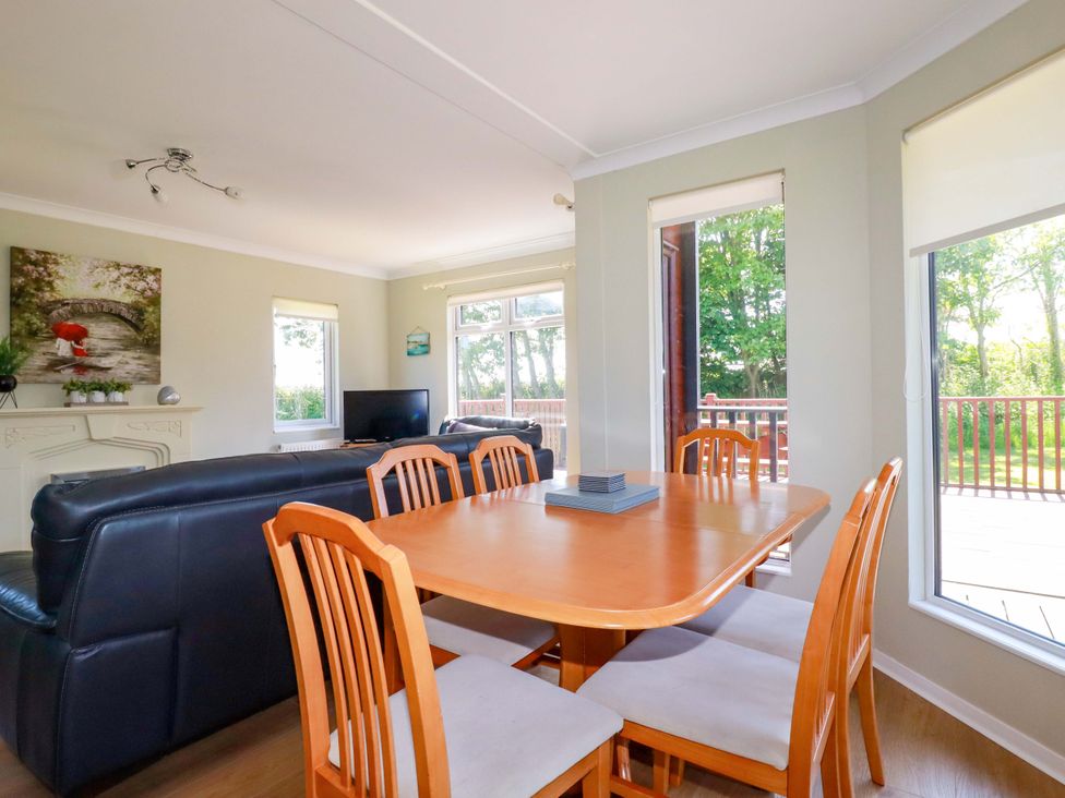 A dining table with six chairs next to a living area with black sofas a TV and windows showing trees outside at Lodge 9- Lamorna Tregolds near St Merryn
