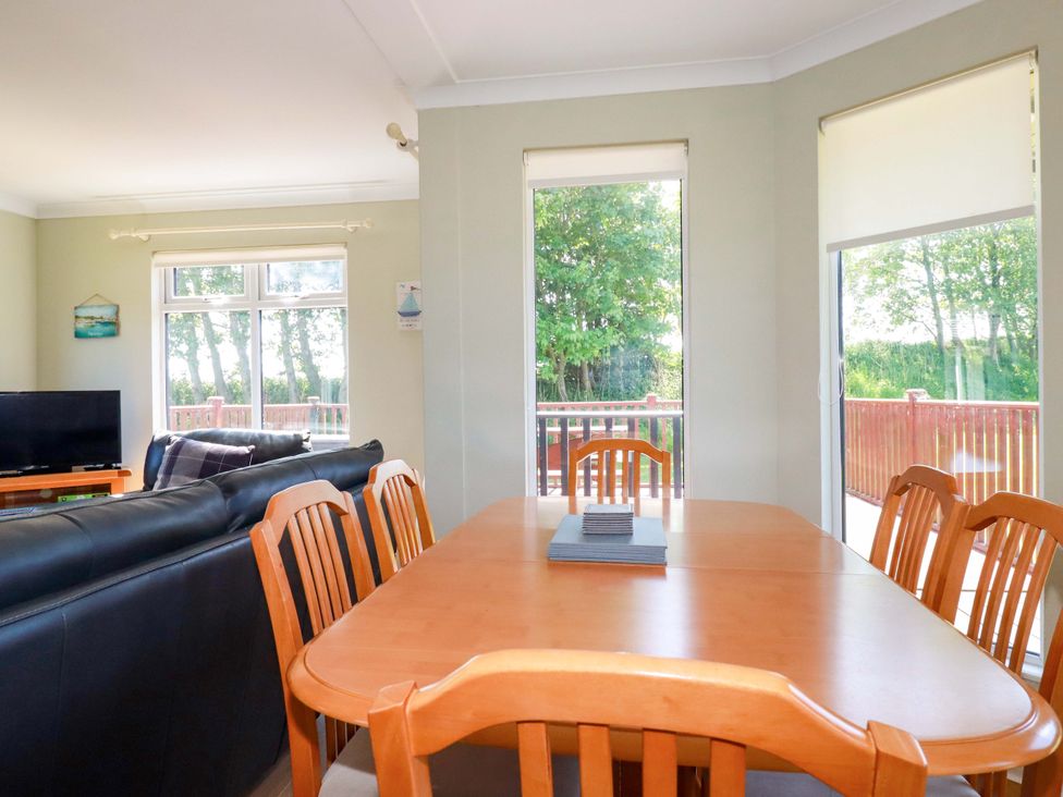 A dining table with wooden chairs in front of large windows and a living room with black sofas and a TV at Lodge 9- Lamorna in Tregolds near St Merryn