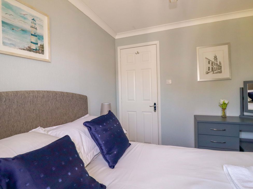 A bedroom with a bed with blue pillows a gray bedside lamp a white door and a dresser with flowers and a mirror at Lodge 9- Lamorna in Tregolds near St Merryn