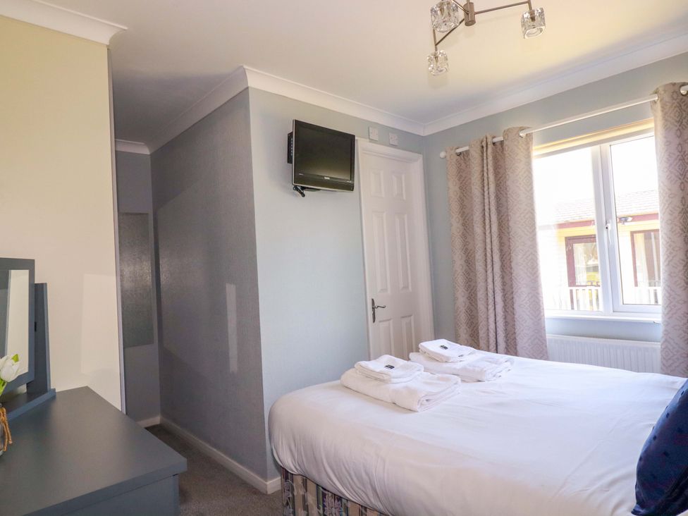 A bedroom with a bed with folded towels a wall-mounted TV a window with curtains and a dresser with a mirror at Lodge 9- Lamorna in Tregolds near St Merryn