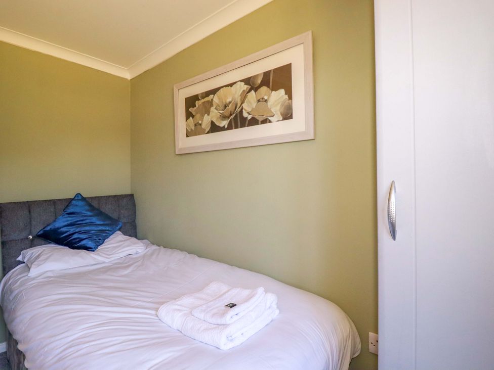A bedroom with a single bed with white linens and blue pillow a white wardrobe and a floral painting on the wall at Lodge 9- Lamorna in Tregolds near St Merryn