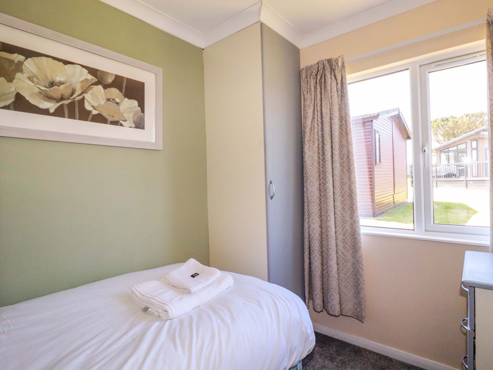 A small bedroom with a single bed with white bedding and towels on top a window with patterned curtains a wardrobe and a painting of flowers on the wall at Lodge 9- Lamorna in Tregolds near St Merryn