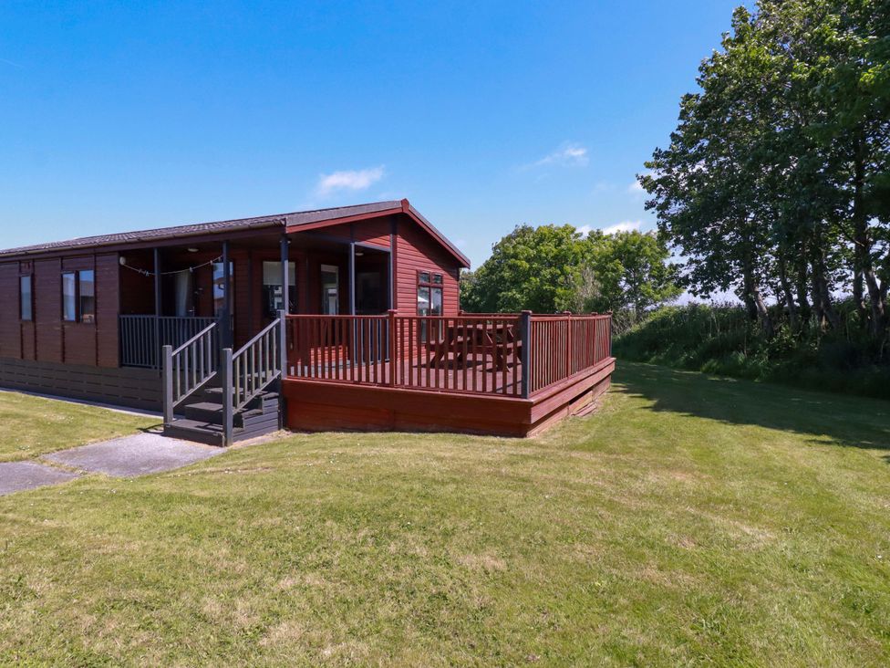 A wooden cabin with a deck and picnic table on a grassy lawn near trees at Lodge 9- Lamorna Tregolds near St Merryn