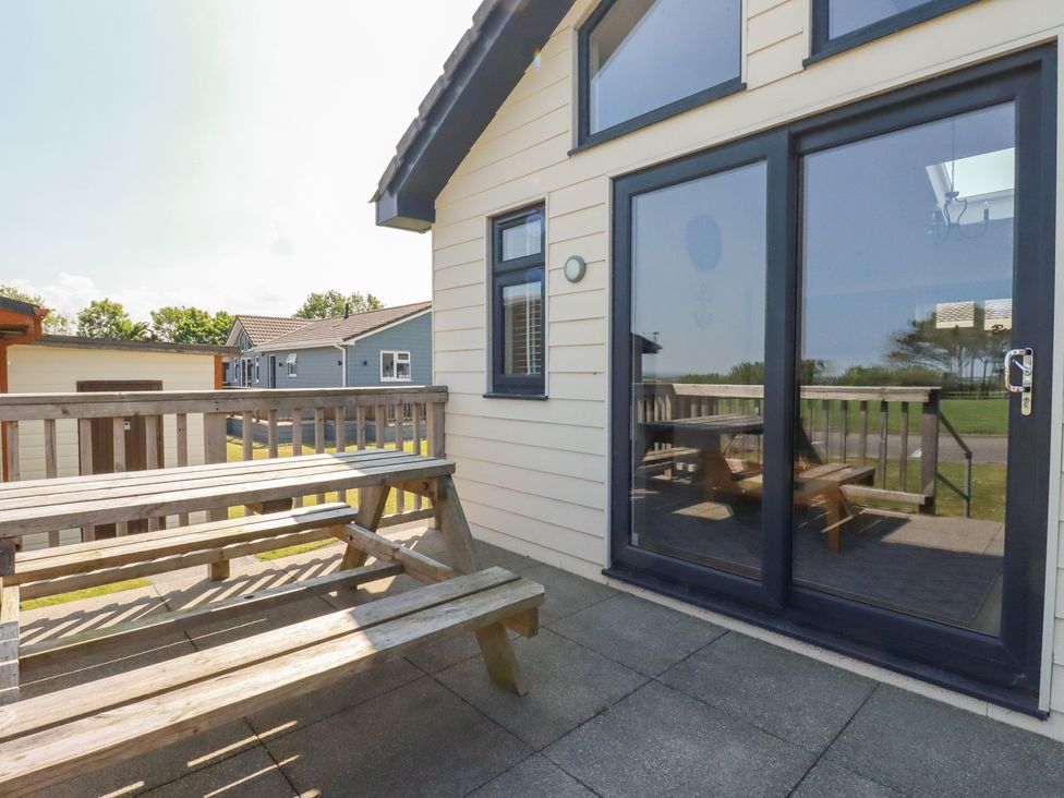 An outdoor patio with a wooden picnic table and benches next to a building with glass sliding doors and a window at Lodge 18 in St Merryn