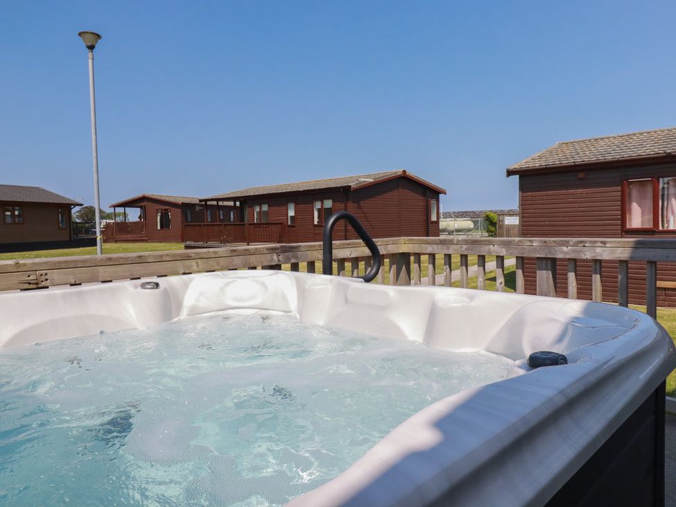 An outdoor hot tub on a deck with wooden cabins and a street lamp in the background at Lodge 18 in St Merryn