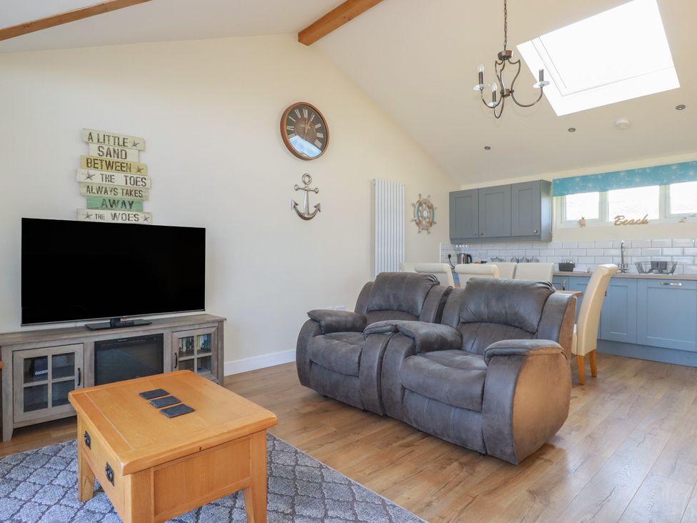 A living area with a grey recliner sofa wooden coffee table television on a stand and a kitchen with blue cabinets at Lodge 18 in St Merryn
