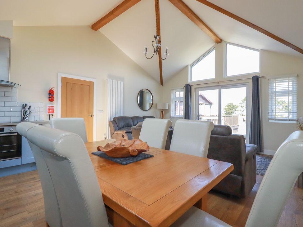 A dining area with a wooden table and leather chairs next to a living room with sofas and large windows at Lodge 18 in St Merryn